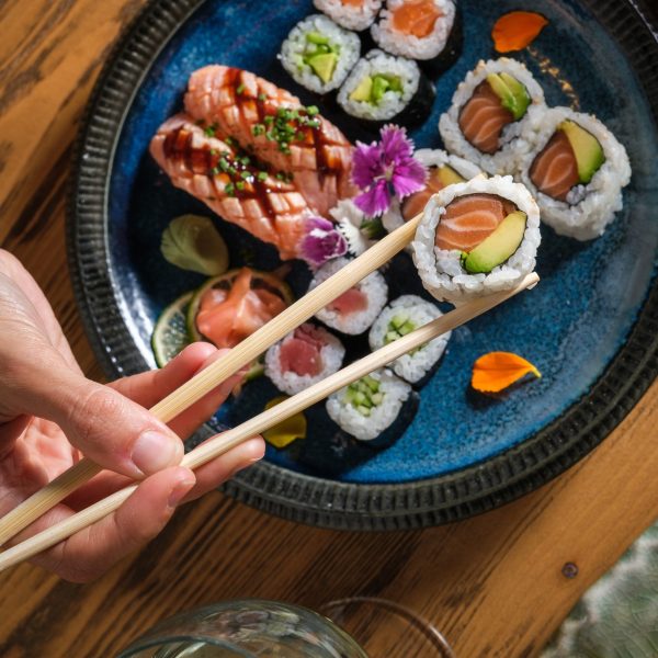 Top view of crop anonymous person with bamboo chopsticks taking tasty sushi roll with raw salmon and avocado from set